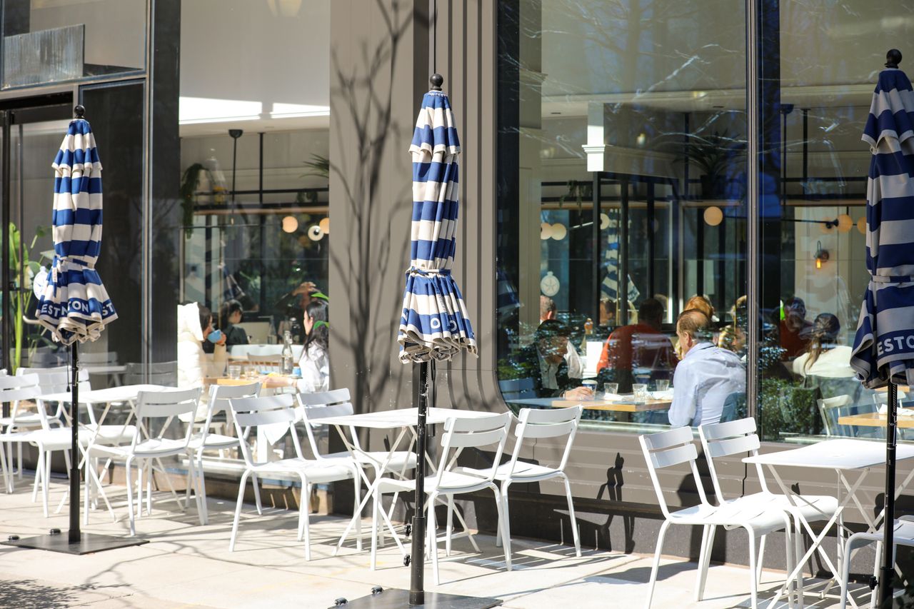 services-01 A sunlit outdoor café scene in Midtown Manhattan, New York, showcasing elegant white chairs and blue striped umbrellas.